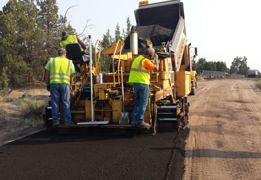 Paving crew riding the machine as hot mix goes down over a freshly compacted stone base